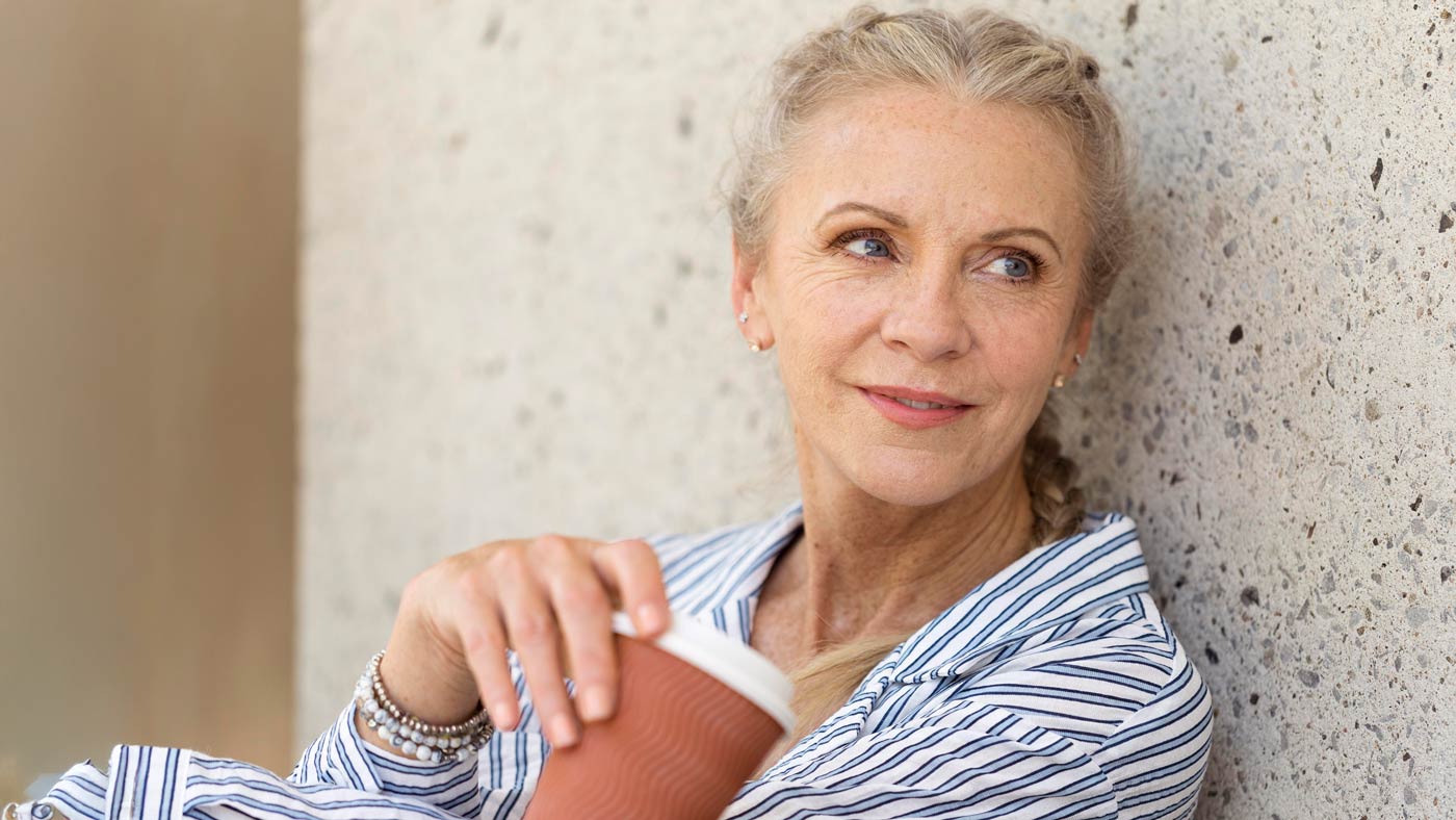 Smiling mature woman with braided hair holding a coffee cup, leaning against a textured wall and wearing a striped shirt—looking relaxed and confident in natural daylight.