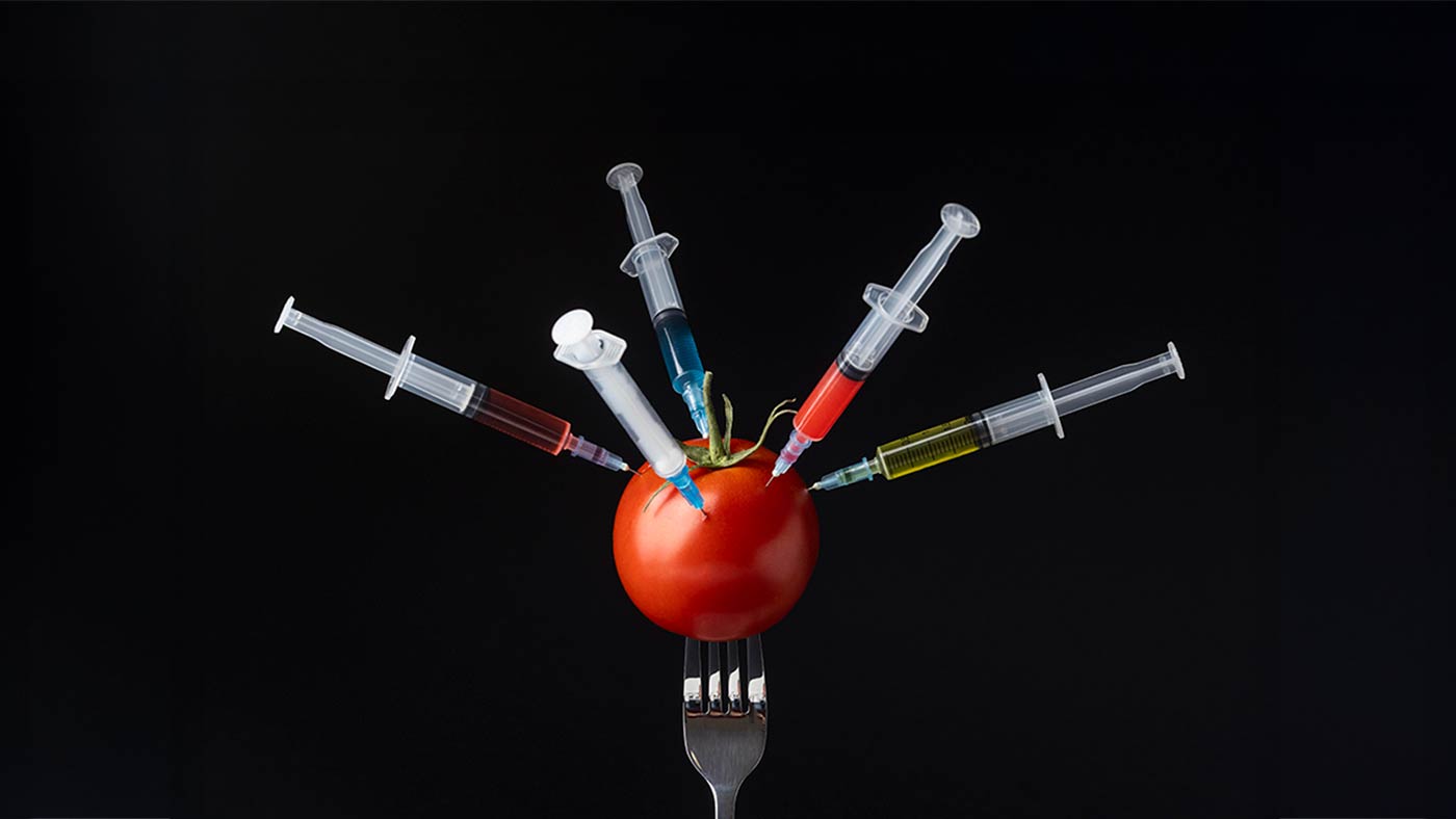 A tomato pierced by multiple syringes filled with colorful liquids, symbolizing concerns about food additives and chemical modifications, set against a black background