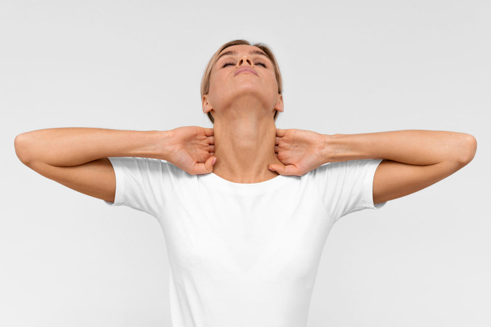 A woman in a white shirt stretching her neck backward while holding the back of her neck with both hands. She appears to be relieving tension or stiffness, set against a plain background, conveying a sense of relaxation or stress relief.