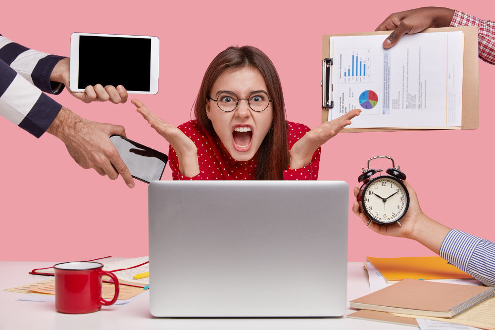 A stressed woman with glasses sitting at a desk in front of a laptop, surrounded by hands holding a tablet, clipboard, clock, and phone, representing multitasking and workplace stress.