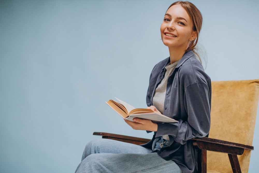 A woman sitting comfortably on a chair, smiling while holding an open book. She is wearing a casual outfit with a light gray top and a blue jacket. The background is simple and light blue, creating a relaxed and inviting atmosphere.