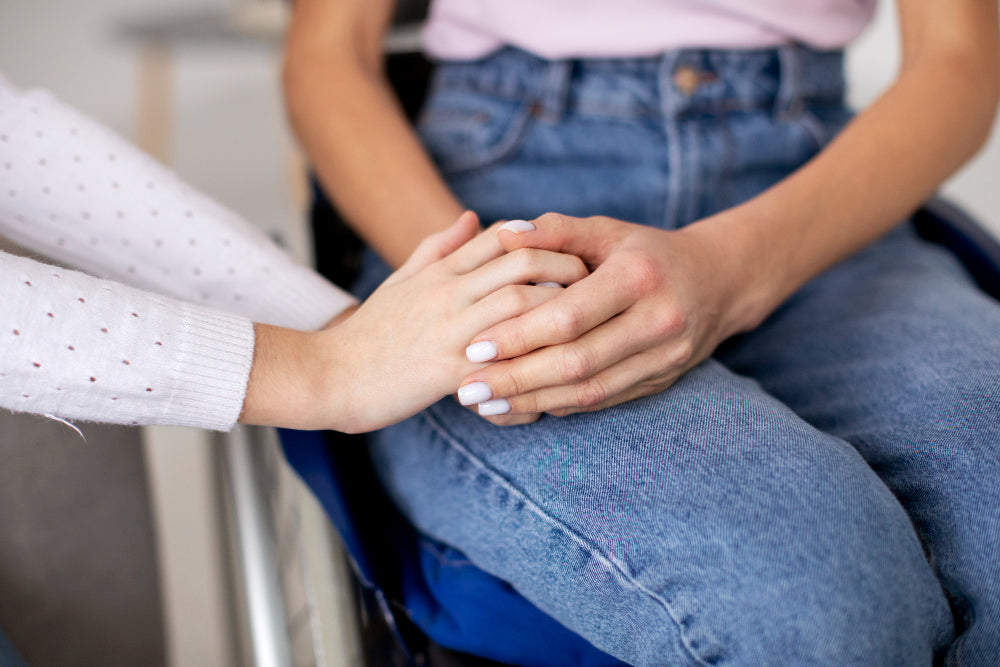 A supportive gesture as one person holds the hand of another seated individual, symbolizing empathy and comfort. The seated person is wearing jeans, and the scene suggests a caring interaction, providing reassurance or emotional support.