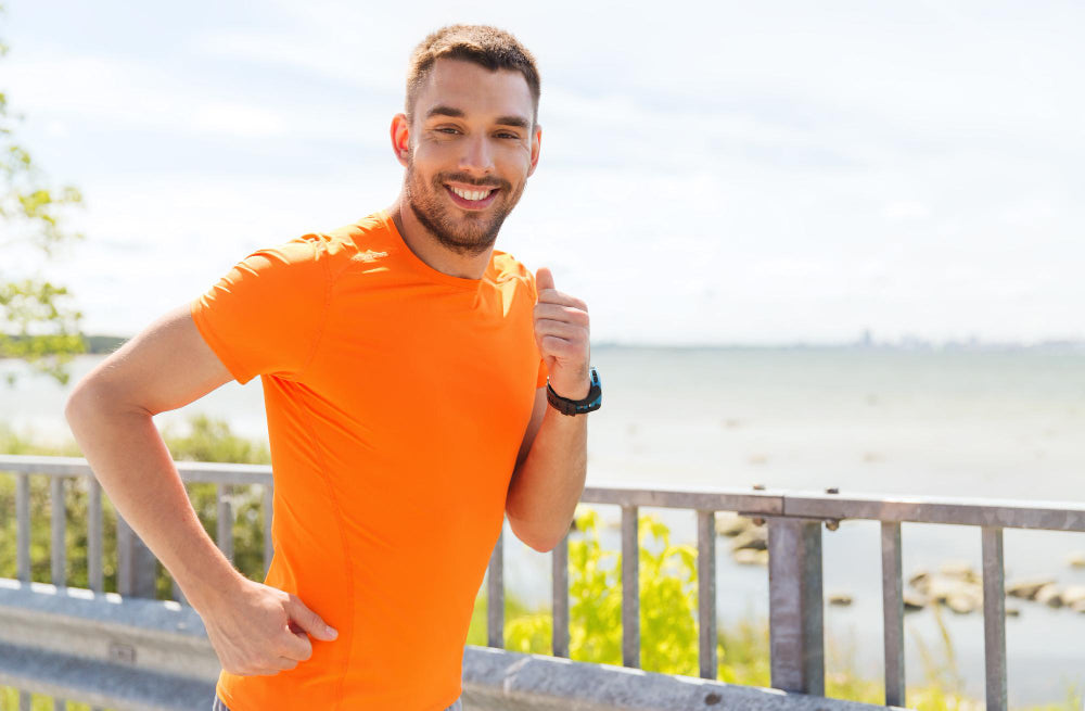 A man in an orange athletic shirt is jogging outdoors near a body of water, smiling at the camera. The image captures a bright, sunny day, with greenery and clear skies in the background, emphasizing an active and healthy lifestyle.