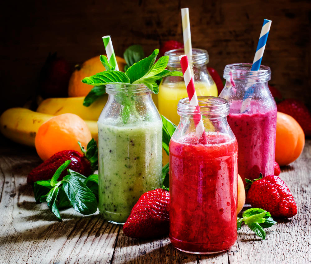 Colorful fruit smoothies in glass bottles with straws, surrounded by fresh fruits and mint leaves on a wooden table.