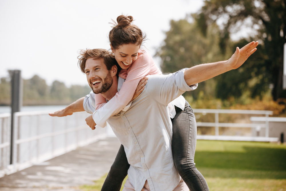 A couple enjoying a playful moment outdoors, with the man giving the woman a piggyback ride. They both have joyful expressions, capturing a sense of fun and lightheartedness against a backdrop of greenery and clear skies.