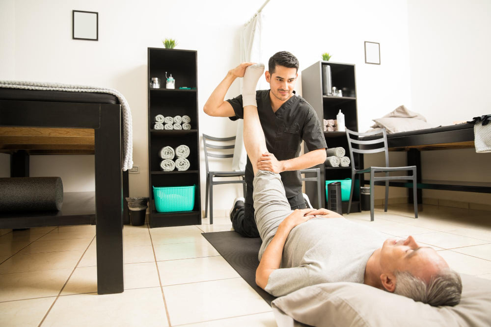 A physical therapist assisting an older adult patient in a rehabilitation session. The patient is lying on a mat while the therapist gently stretches his leg. 