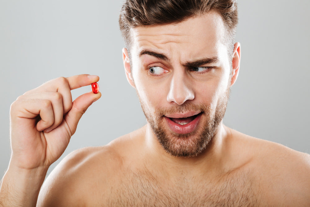 A man with a curious and slightly skeptical expression holding a red capsule between his fingers, looking at it intently. He is shirtless and set against a neutral background.