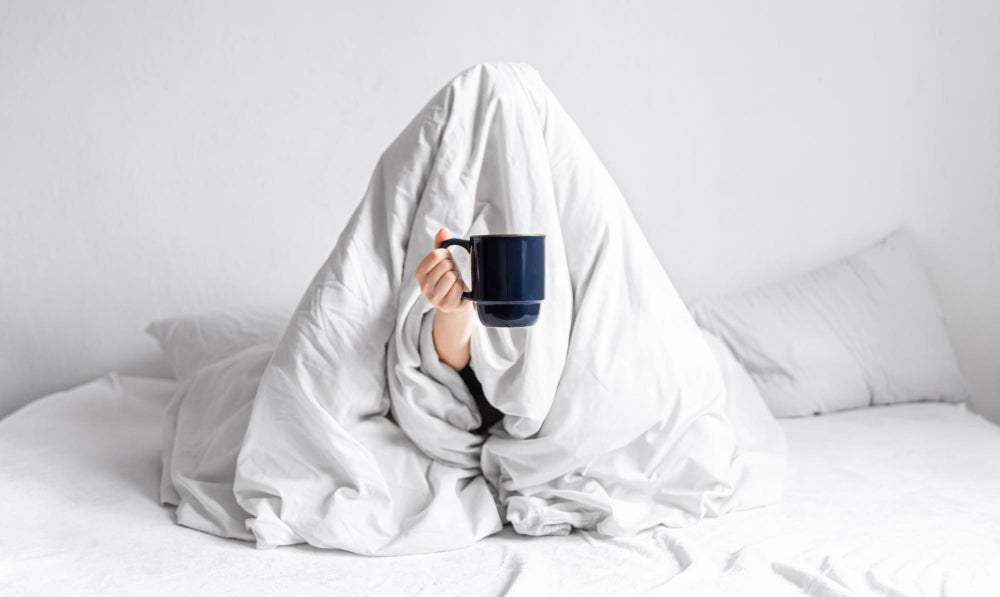 A person wrapped in a white blanket, holding a large coffee mug, with only their arm visible from under the blanket. The image suggests a cozy, sleepy morning, emphasizing the need for comfort and rest.