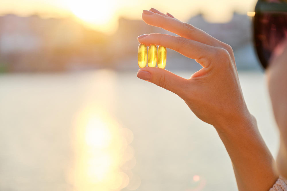 A hand holding two golden vitamin D capsules against a bright sunset, symbolizing the connection between sunlight and vitamin D supplementation.