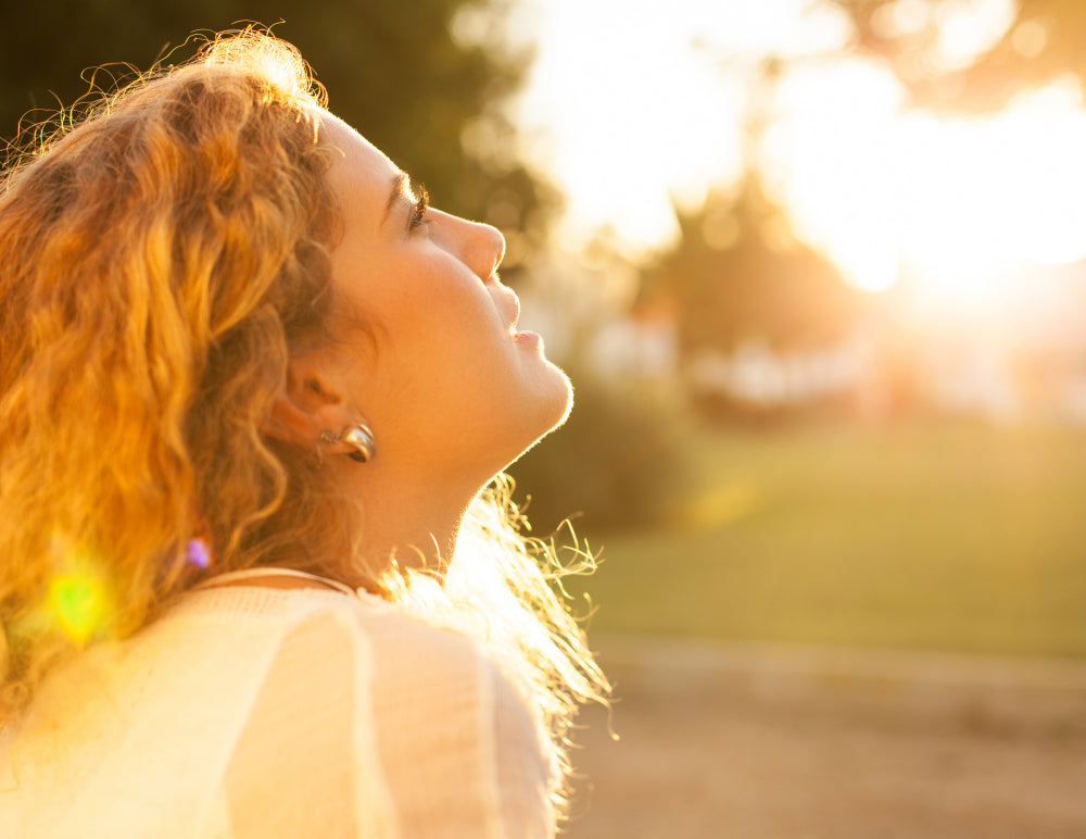 A woman with curly blonde hair looks upward with her face bathed in warm sunlight, reflecting a peaceful and serene expression as the sun sets in the background.